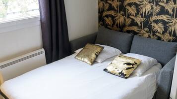 Una cama con ropa blanca y dos almohadas de diseño dorado y negro, un cabecero gris y una ventana con cortina.