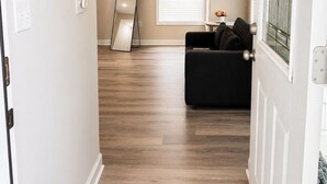 A hallway with a wooden floor, a black couch, a large mirror, and a window with blinds.