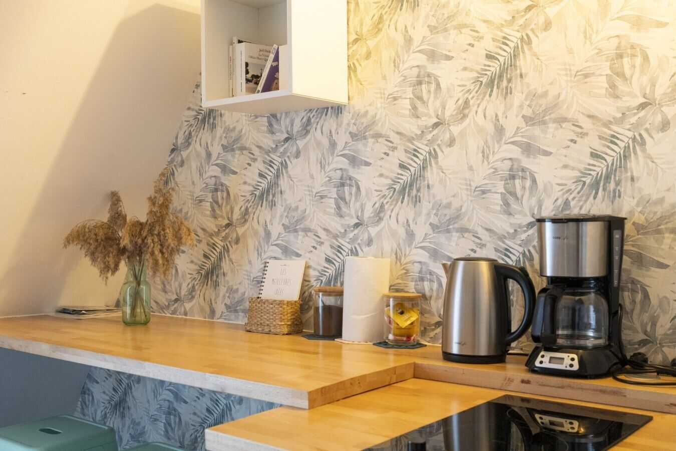 A kitchen with a coffee maker, a kettle, a vase with dried flowers, and a shelf with books.
