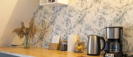 A kitchen with a coffee maker, a kettle, a vase with dried flowers, and a shelf with books.