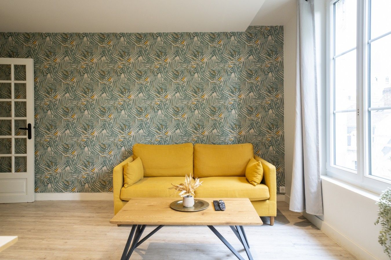 A room with a patterned wallpaper, a yellow sofa, a wooden coffee table, and a window with a view.