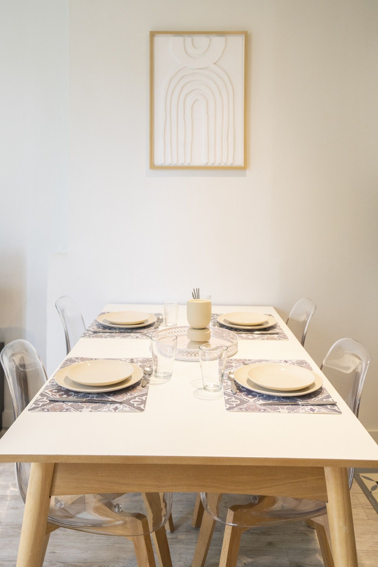 A dining table set for six with clear glassware and plates, a centerpiece, and a framed artwork on the wall.