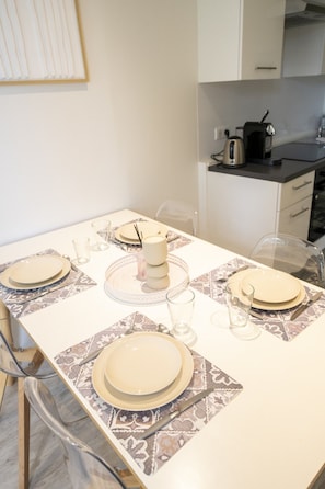 Mesa de comedor dispuesta con platos, vasos y cubiertos, con una zona de cocina al fondo.