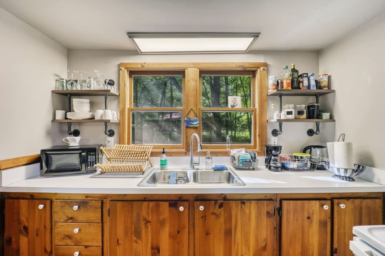 A kitchen with a large window, wooden cabinets, a microwave, and various kitchen items on shelves.