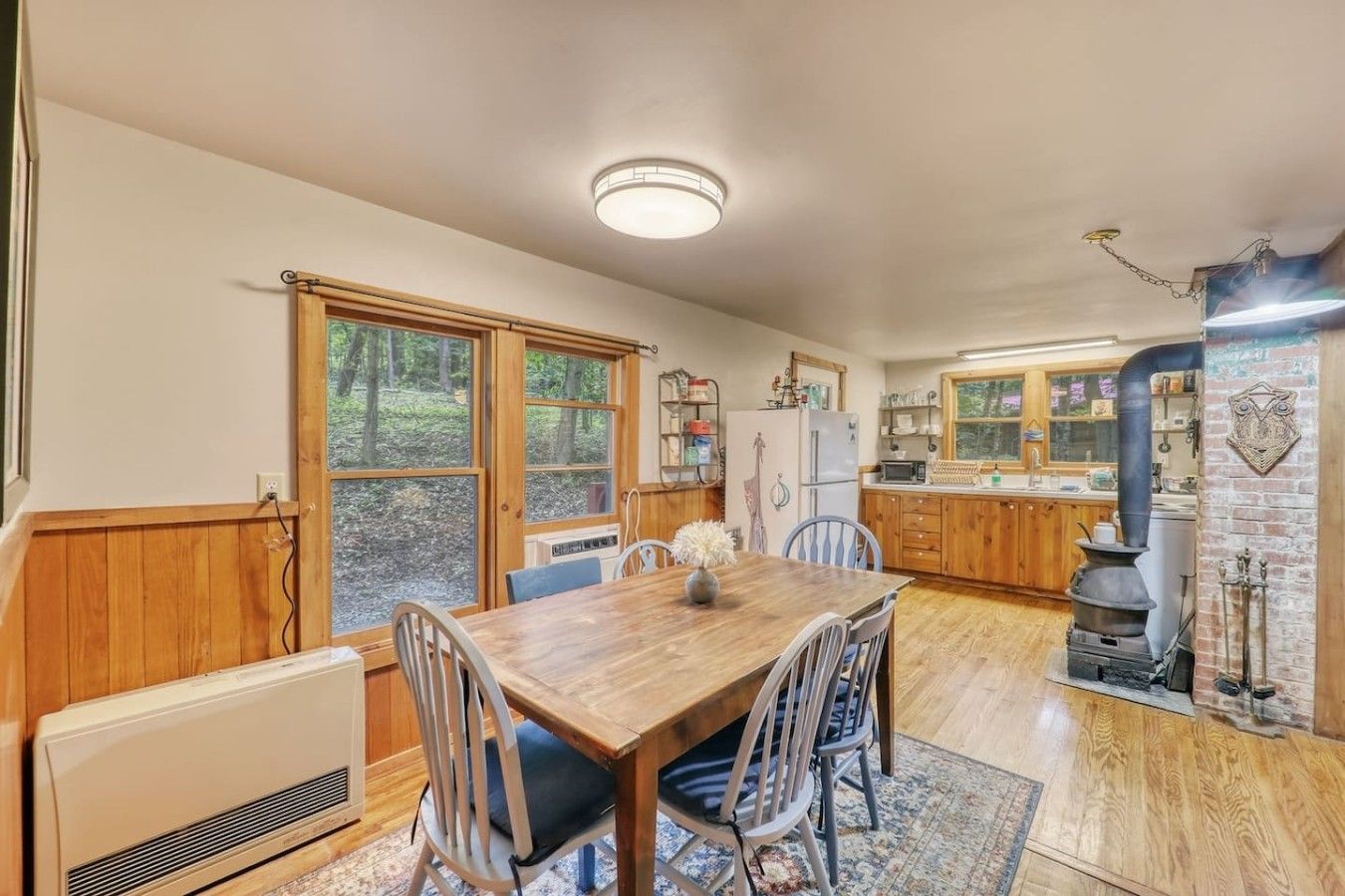 Une cuisine chaleureuse avec des meubles de rangement en bois, une table à manger avec des chaises, une grande fenêtre et un poêle à bois.