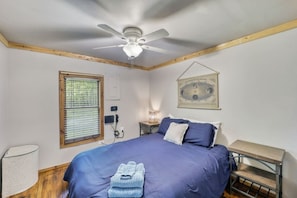 Une chambre à coucher comprenant un lit, un ventilateur de plafond, une fenêtre, un luminaire fixé au mur et une étagère en bois.