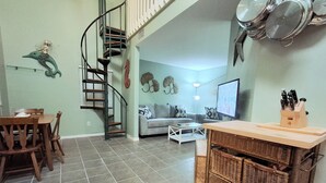 Une cuisine avec un coin repas, un escalier en colimaçon et un salon comprenant un canapé et un téléviseur.