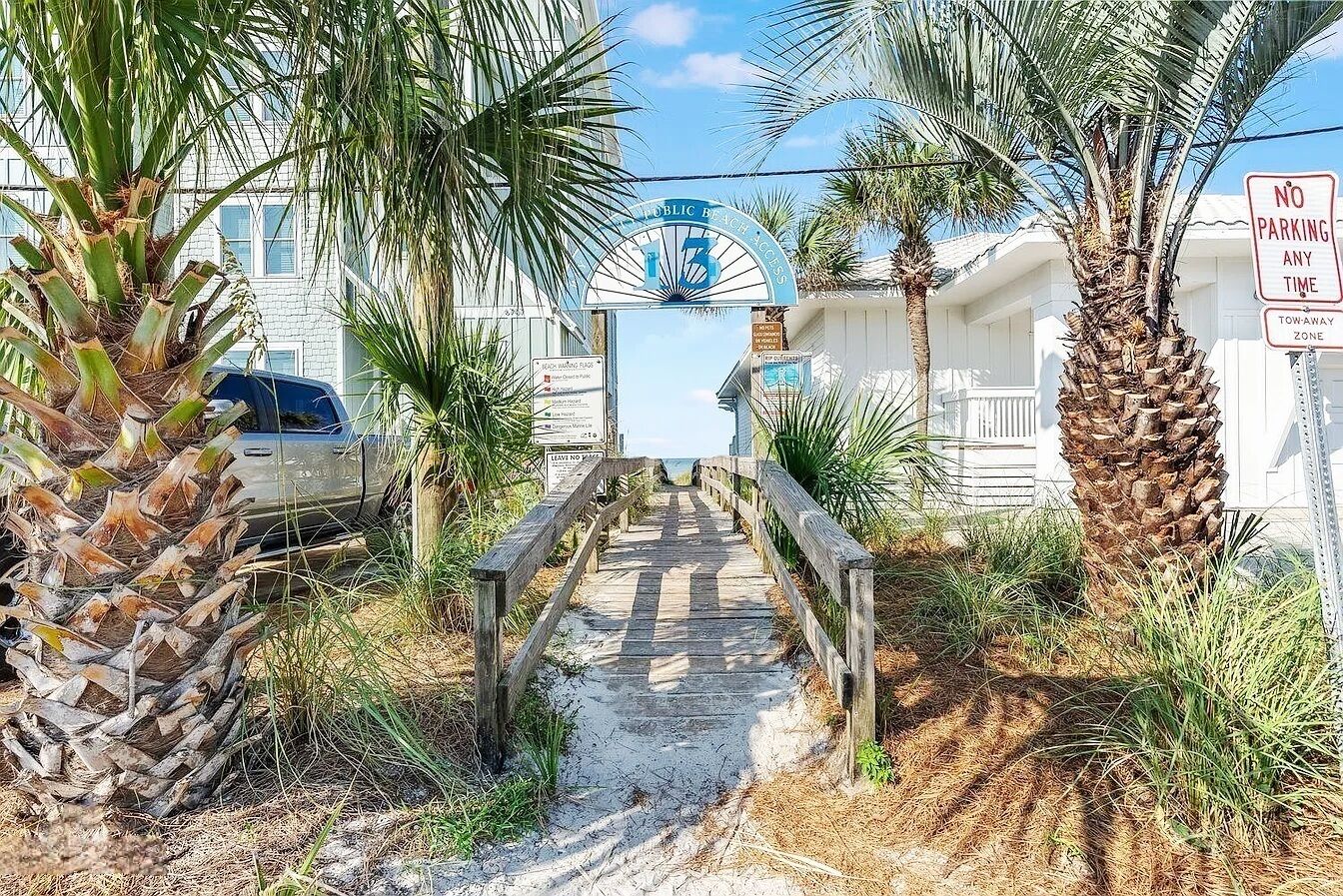 A wooden boardwalk leading to a beach entrance, surrounded by palm trees and a sign indicating no parking at any time.