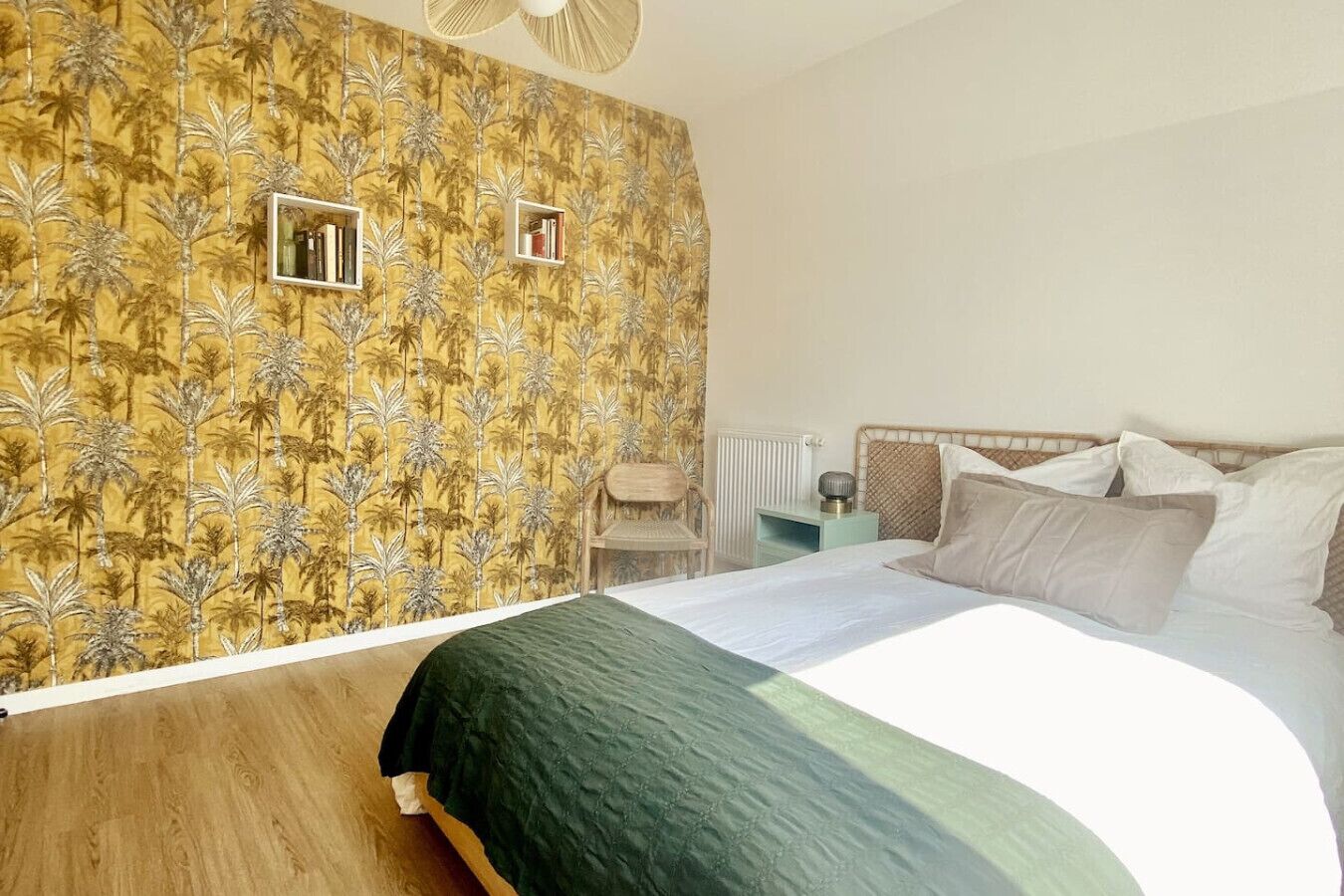 Une chambre à coucher comprenant un lit, une table de chevet, un radiateur et un mur recouvert d’un papier peint à motifs tropicaux.