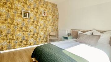 Une chambre à coucher comprenant un lit, une table de chevet, un radiateur et un mur recouvert d’un papier peint à motifs tropicaux.