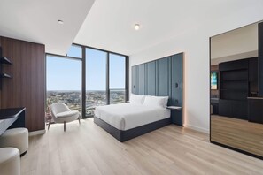 Ein modernes Schlafzimmer mit einem großen Bett, einem Schreibtisch und einem Sessel, mit Blick auf die Stadt durch große Fenster.