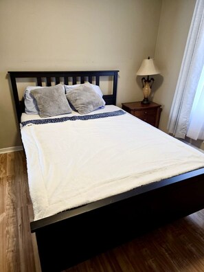 Una cama con edredón blanco, dos almohadas, colcha estampada, cabecero de madera, mesita de noche con lámpara y ventana con cortinas.