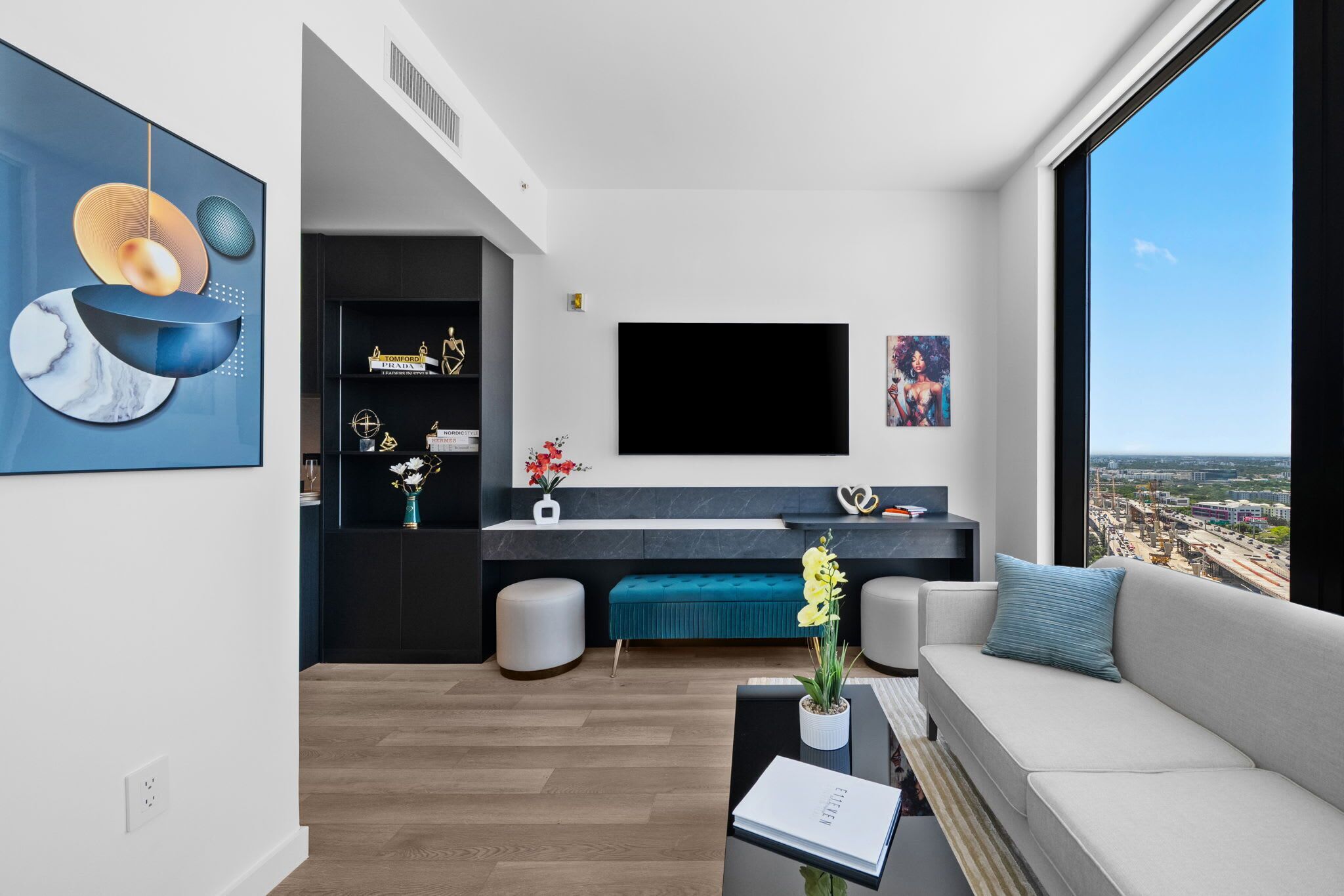 A modern living room with a sofa, coffee table, and TV. There is a large window with a city view.