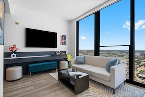 A modern living room with a large TV, a sofa, a coffee table, and a view of the city through large windows. - 32nd Floor Suite w/ Rooftop Pool & Skyline Views (Miami)