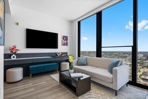 A modern living room with a large TV, a sofa, a coffee table, and a view of the city through large windows. - E11EVEN Superior Suite with Elite Amenities (Miami)