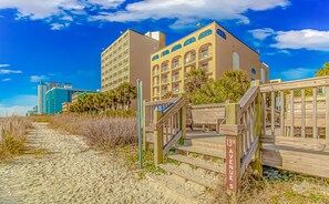 A beachfront hotel with a wooden boardwalk leading to the entrance, a clear blue sky, and a building with multiple balconies. - Sea Mist 50810 Ocean View Escape (Myrtle Beach)