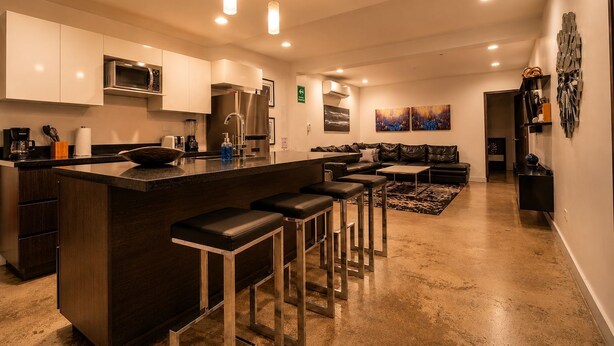 Une cuisine moderne avec un îlot central, des tabourets de bar, un coin repas avec un canapé et un salon agrémenté d’un grand tapis.
