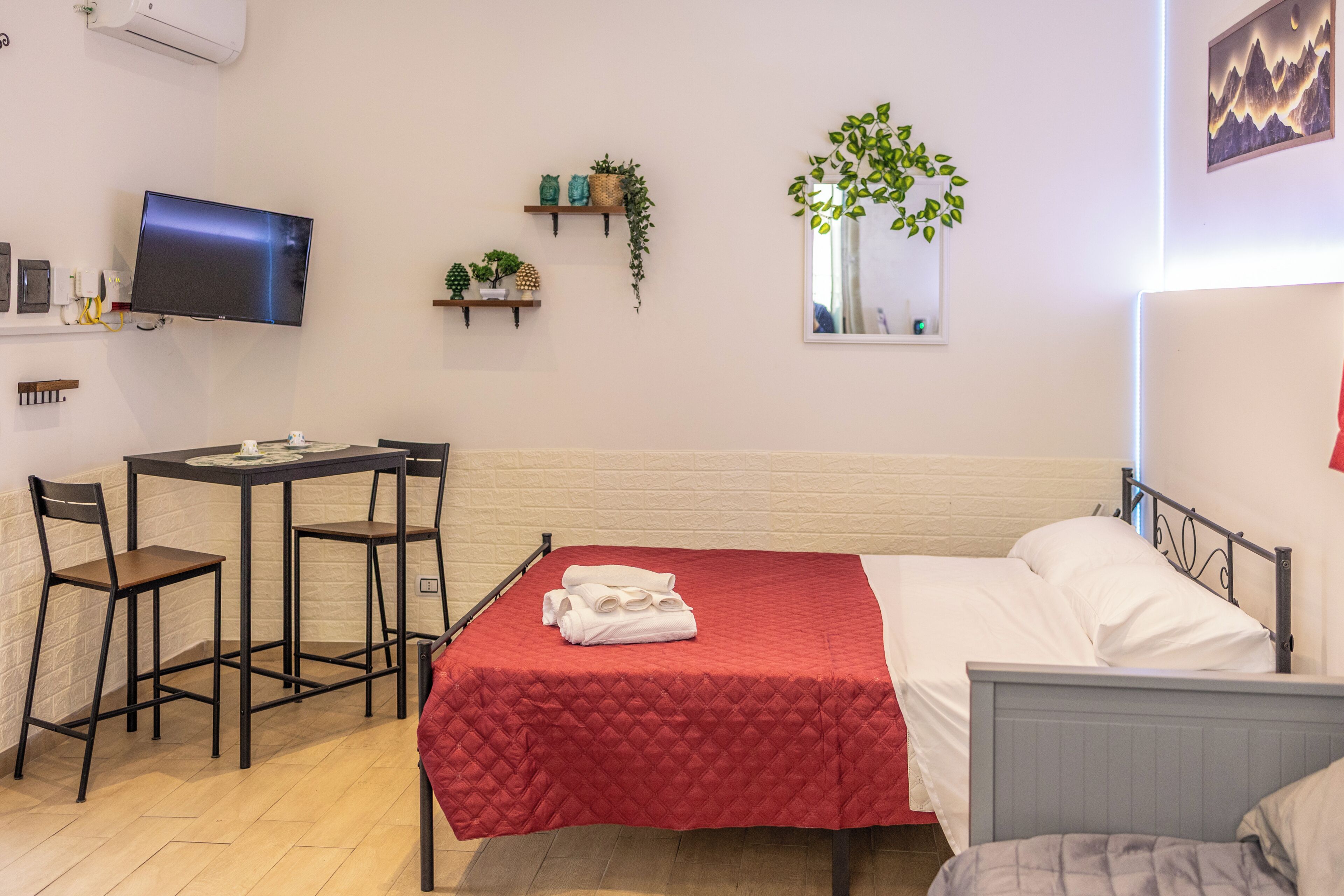 A hotel room with a bed, a desk with chairs, a TV, a wall-mounted shelf with plants, and a window with a view.
