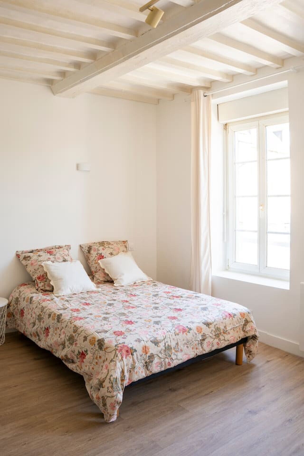 A bedroom with a wooden floor, a bed with floral bedding, a window with curtains, and a ceiling with exposed beams.