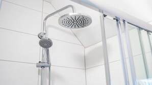 Une douche équipée d’un pommeau amovible et d’une pomme de douche fixe, dans une salle de bain moderne aux carreaux blancs et dotée d’une paroi en verre.