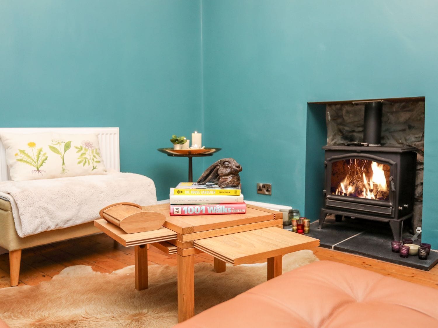 Una habitación acogedora con una pared verde azulado, una cama con una colcha blanca, una pequeña mesa de madera, una estantería llena de libros, una chimenea con fuego encendido y una planta pequeña sobre una mesita de noche.