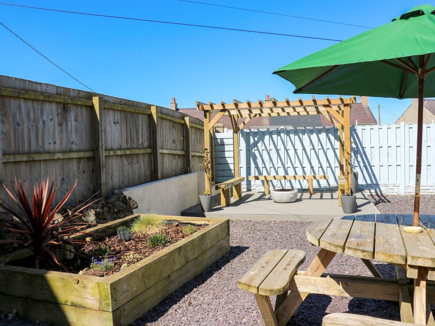 A wooden deck with a pergola, a raised garden bed with plants, and a picnic table with an umbrella.