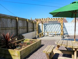 Una terraza de madera con un pérgola, un jardín elevado con plantas y una mesa de picnic con sombrilla.