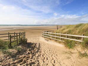 Un sendero de arena con barandas de madera que conduce a una playa con dunas y cielo despejado.
