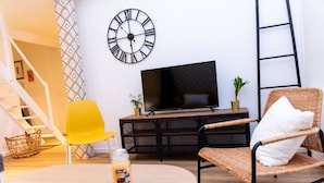 Een moderne woonkamer met een houten tafel, een gele stoel, een rieten stoel met een wit kussen, een zwarte klok, een flatscreen televisie en een trap.