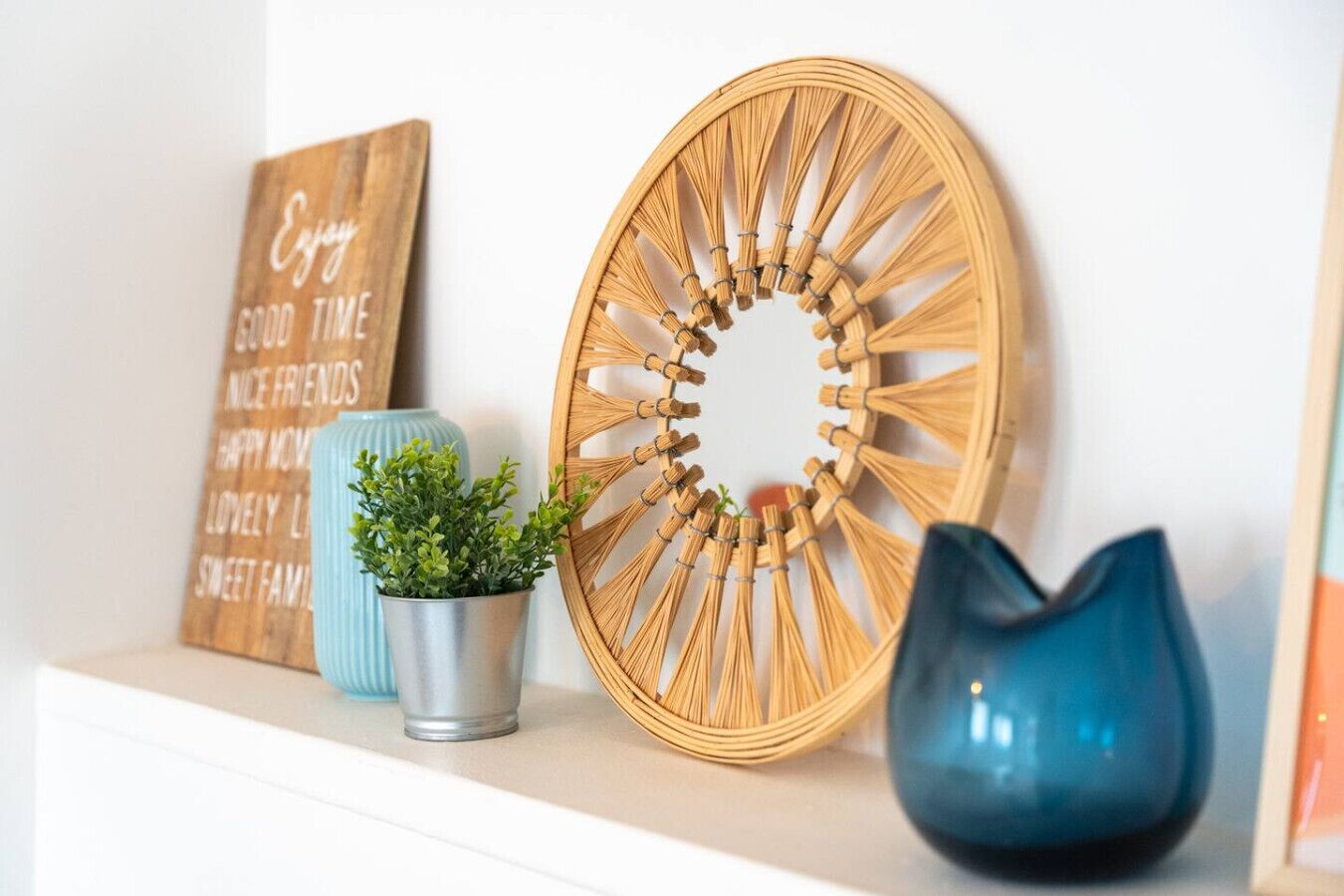 A wall with a wooden sign, a woven wall hanging, a potted plant, and a blue vase.