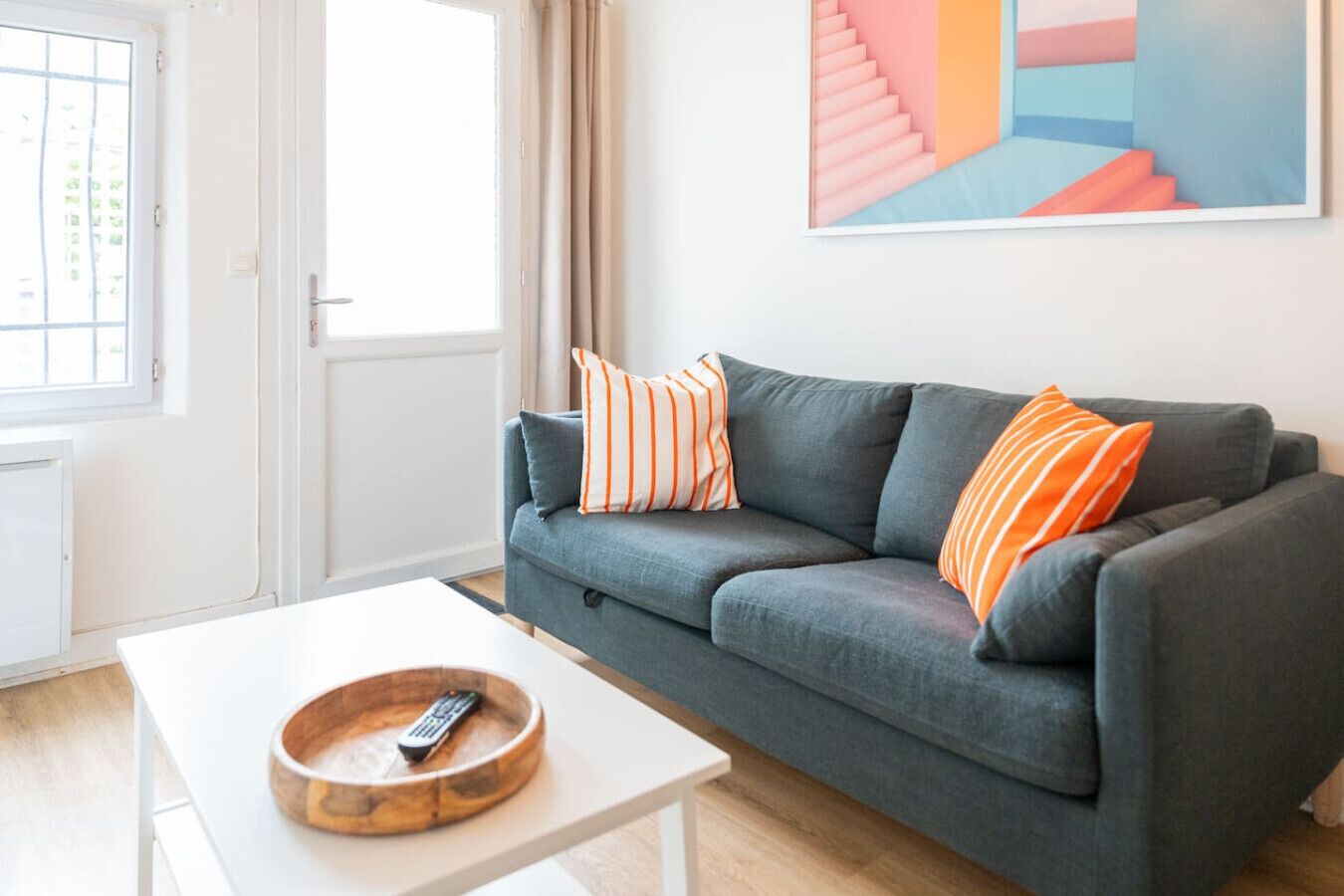 Un salon avec un canapé gris, des coussins à rayures, une table basse blanche et une télécommande.