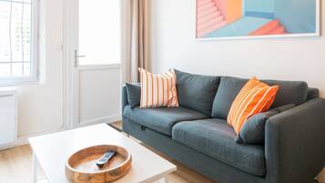 Un salon avec un canapé gris, des coussins à rayures, une table basse blanche et une télécommande.