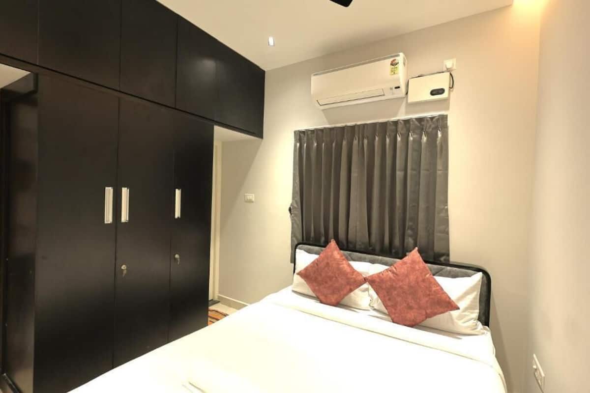 Quarto com cama, guarda-roupa escuro, aparelho de ar condicionado e janela com cortinas.