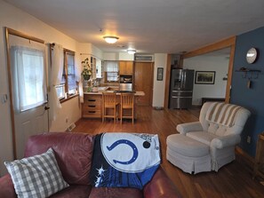 Ein gemütliches Wohnzimmer mit einem roten Ledersofa, einem weißen Sessel und einer Decke mit einer Zeichentrickfigur. Im Hintergrund ist eine Küche mit Holzschränken und einem Kühlschrank zu sehen.