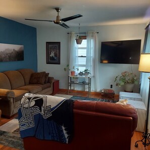 Ein Wohnzimmer mit einer Couch, einem Fernseher, einem Deckenventilator und einem Fenster mit Vorhängen.