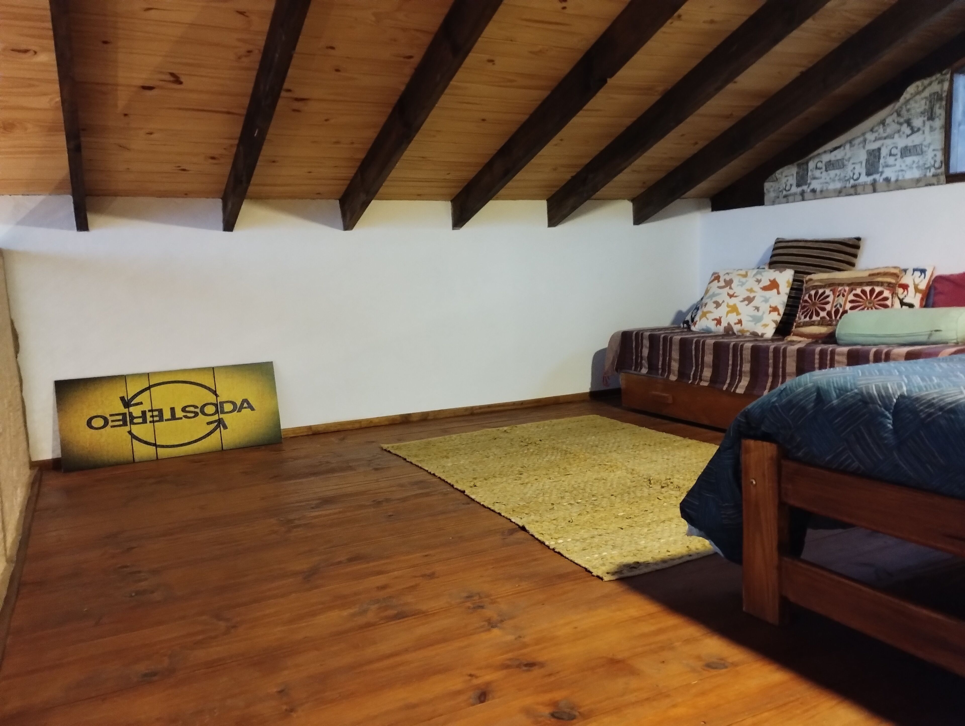 Een kamer met een houten plafond, een bed met beddengoed met een patroon, een geweven vloerkleed en een bord met tekst.