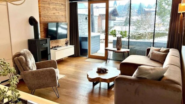 Een moderne woonkamer met een bank, fauteuil, televisie, open haard en grote ramen met uitzicht.