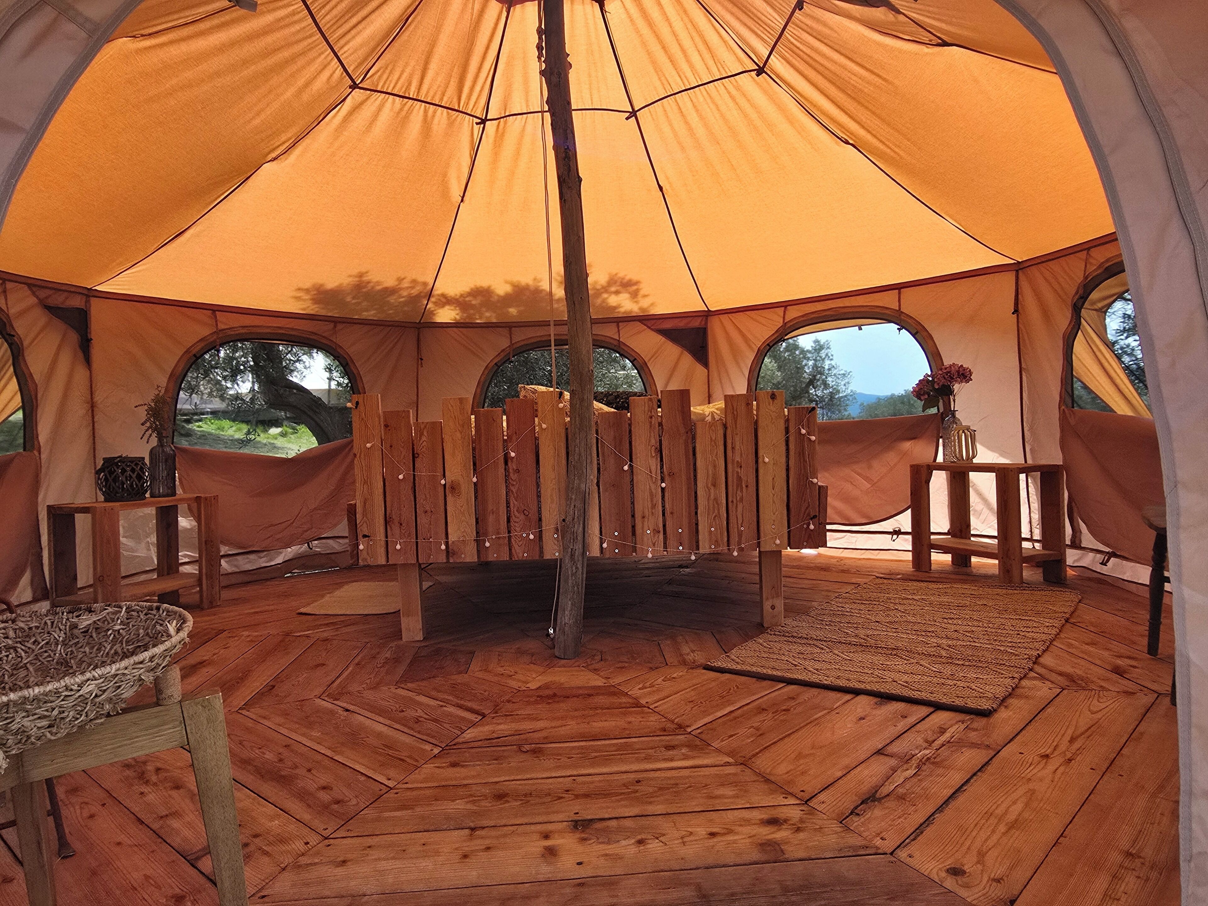 Interior de uma tenda com piso de madeira, um banco, uma mesa pequena com um vaso e vista para o exterior pelas janelas.