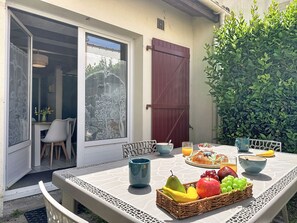 A dining table with breakfast items, a wicker fruit basket, and a view of a house through a glass door. - Port Saint-Jacques by Interhome (La Bernerie-en Retz)
