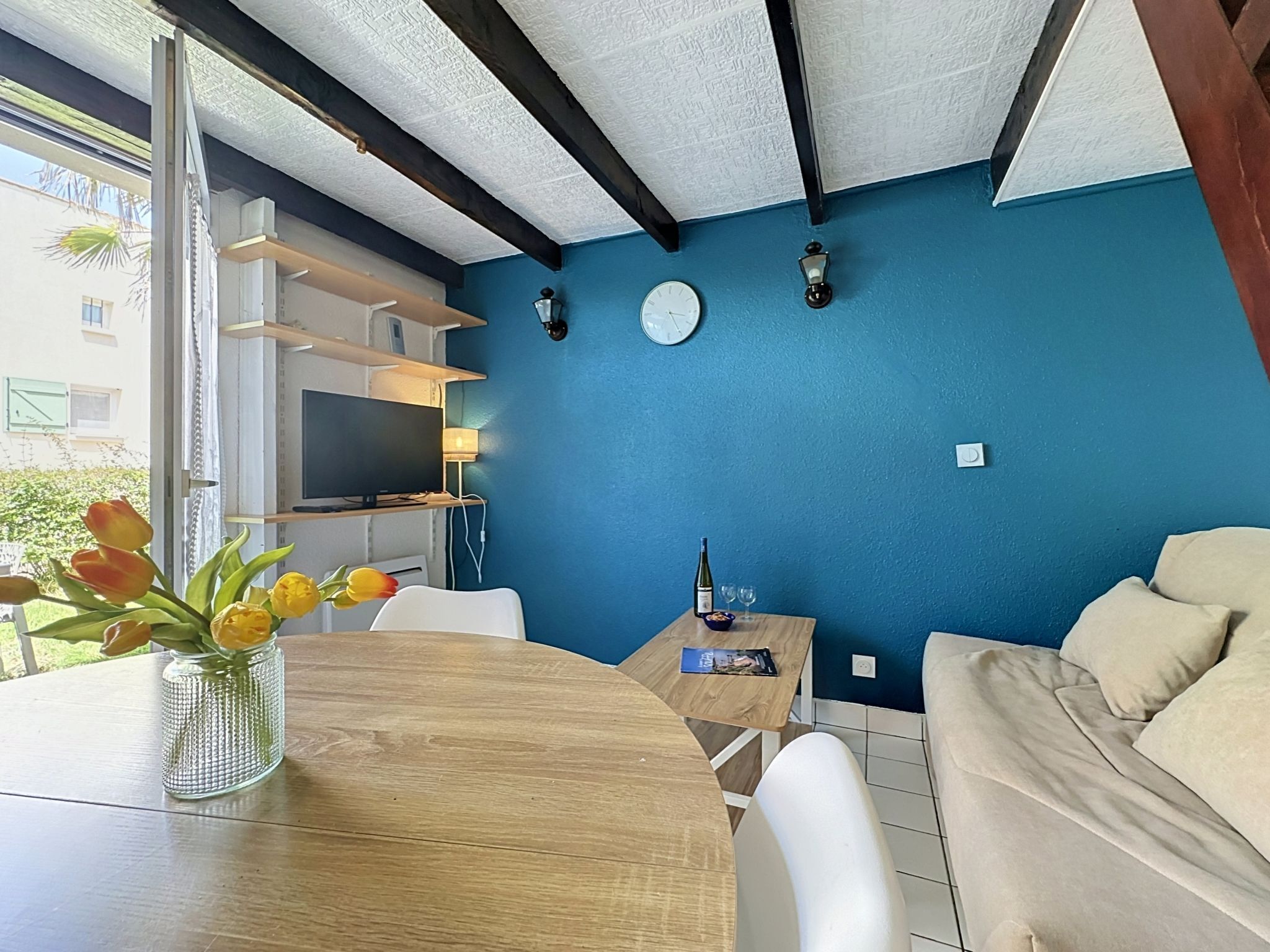 A room with a dining table, chairs, a sofa, a TV, a clock, and a window with a view of buildings.