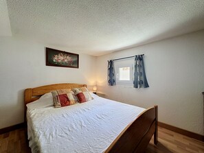 A bedroom with a wooden bed, a nightstand, a window with curtains, and a painting on the wall. - Port Saint-Jacques by Interhome (La Bernerie-en Retz)