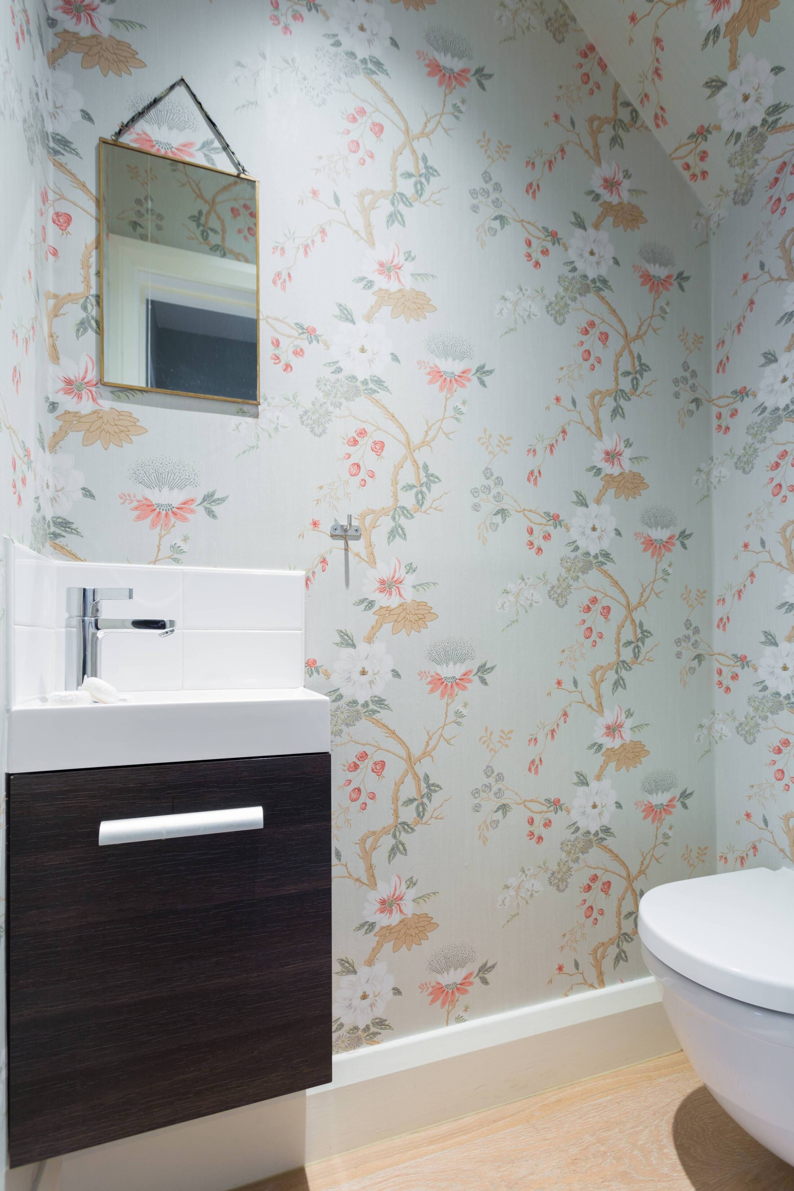 Un bany amb paper pintat floral, un lavabo blanc, un armari de fusta fosca i un vàter blanc.