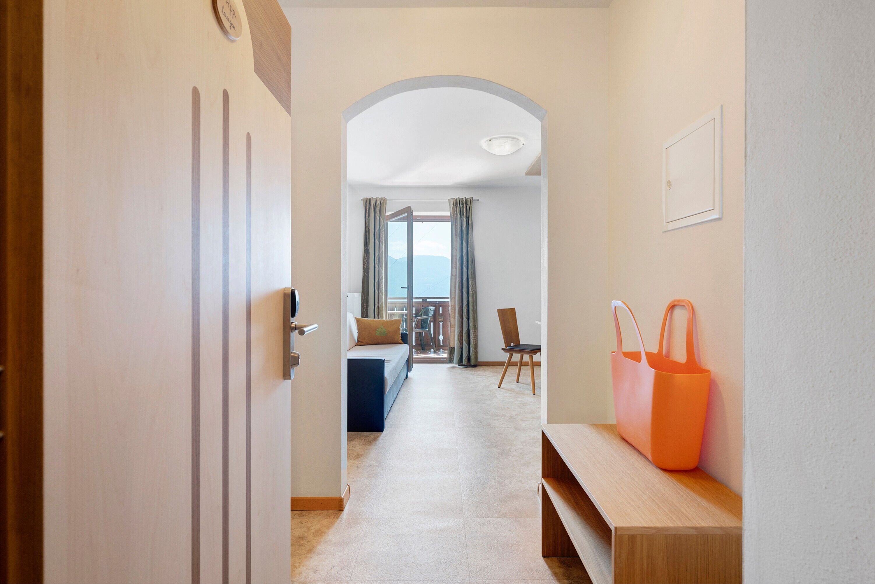 A hallway leading to a balcony with a sofa and curtains, a wooden bench, and a wall-mounted orange bag.