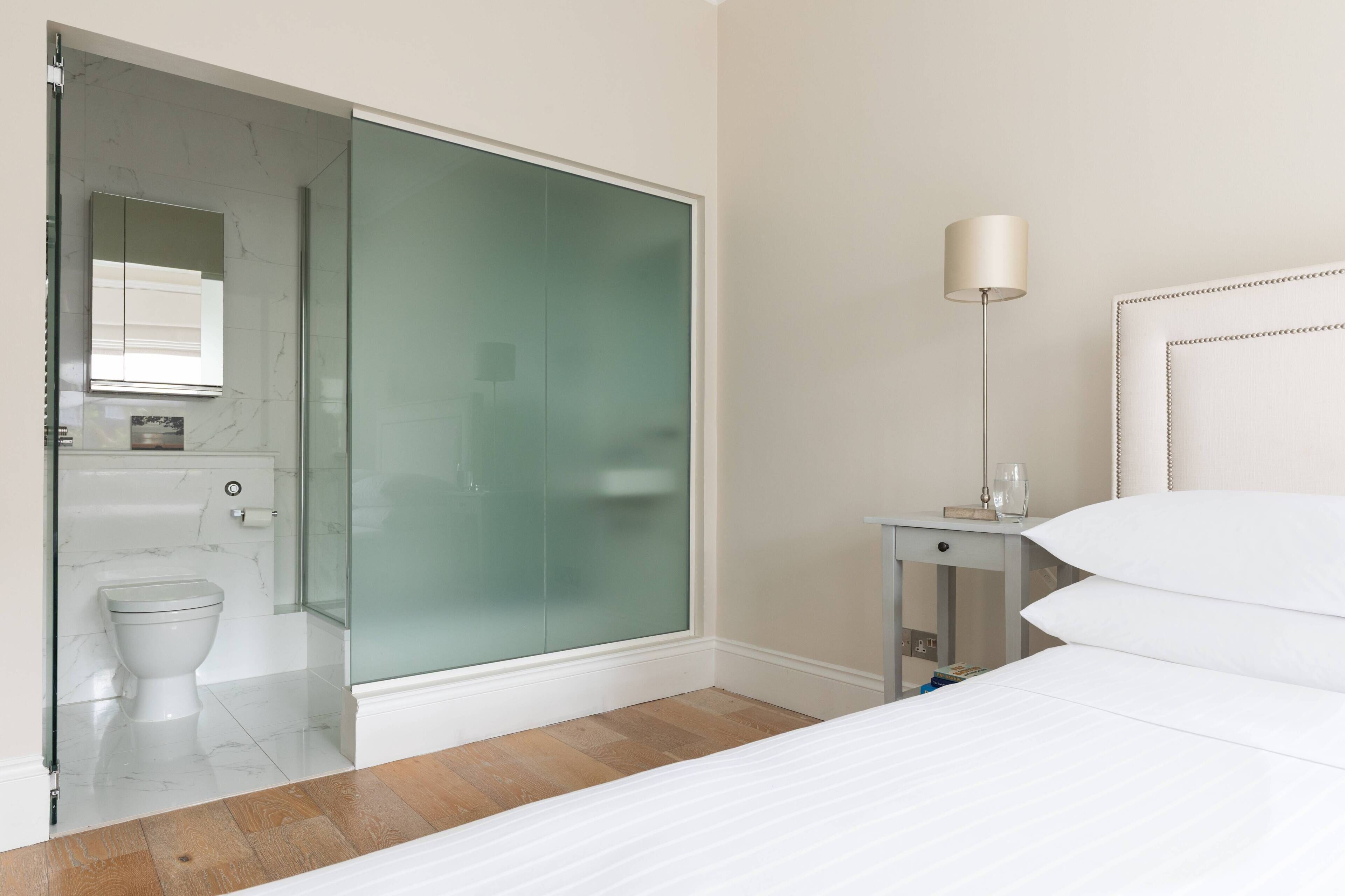 A hotel room with a glass shower door, a bedside table with a lamp, and a bed with white bedding.