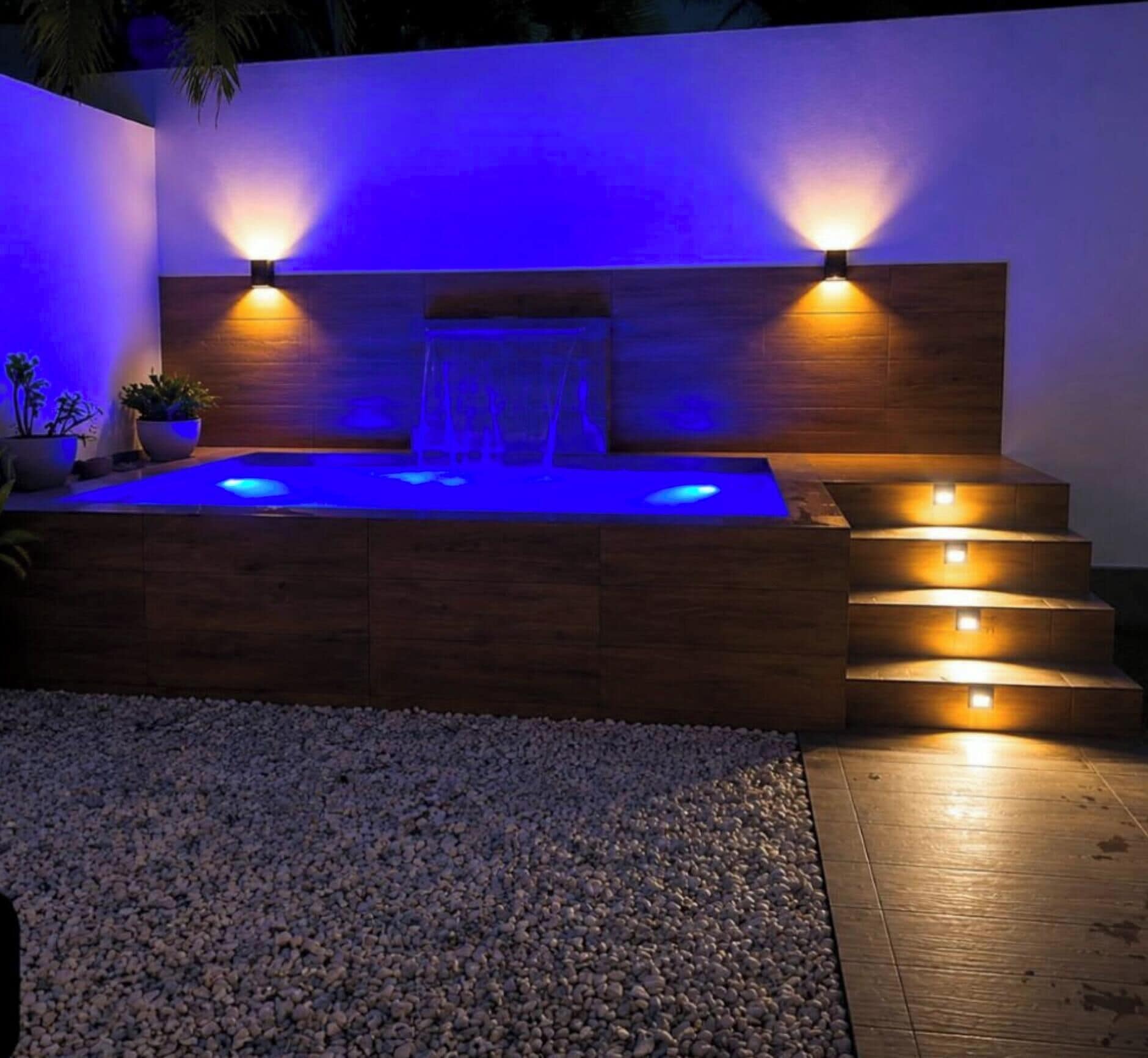 Een hottub met een houten bank, twee wandlampen en een waterval.