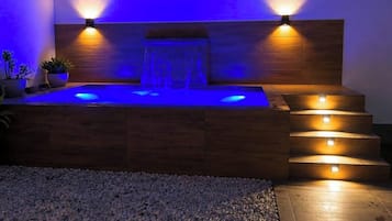 Een hottub met een houten bank, twee wandlampen en een waterval.