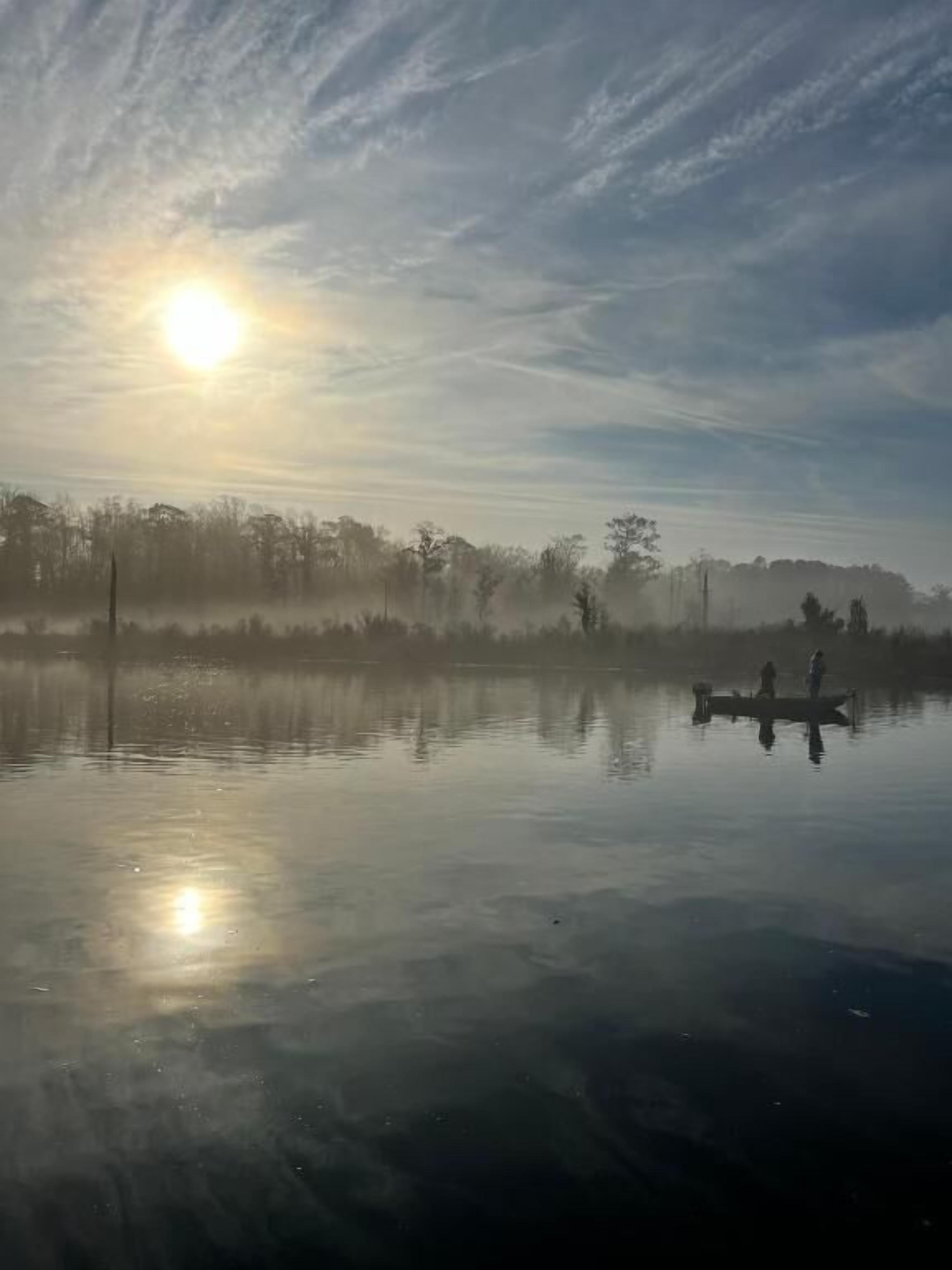 Un lago tranquilo con un bote y gente, rodeado de árboles y un cielo despejado.