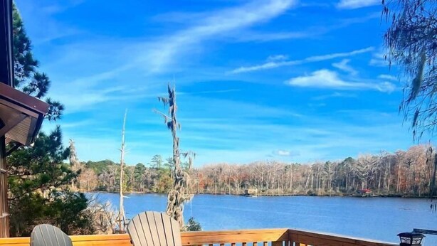 Una terraza de madera con sillas Adirondack y una mesa, con vistas a un lago y árboles al fondo.