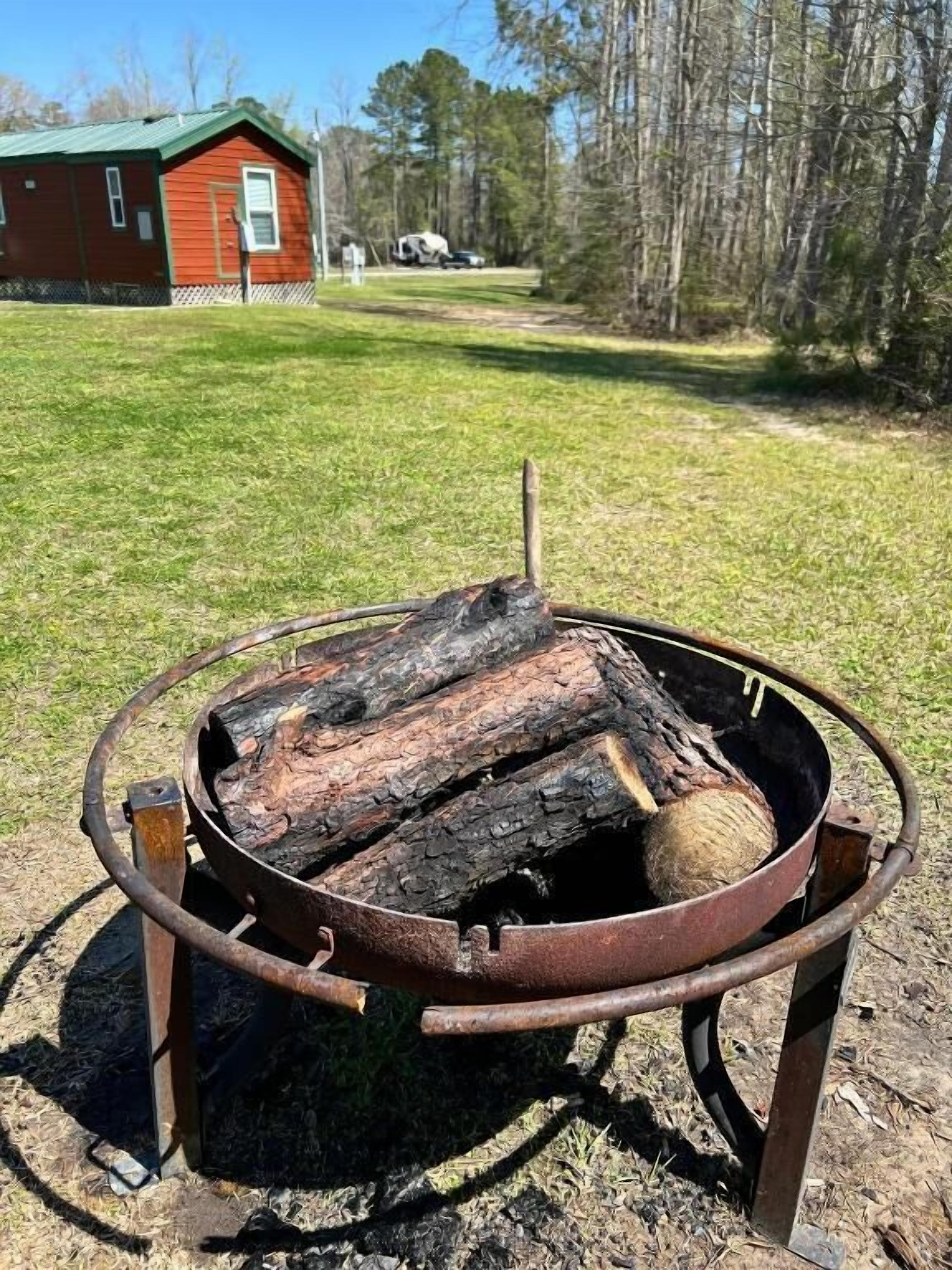 A metal fire pit with logs, a red building in the background, and a grassy area.