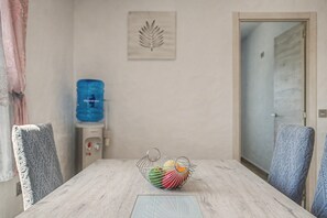 Une table à manger avec un panier de fruits, des chaises, un distributeur d’eau et une porte.